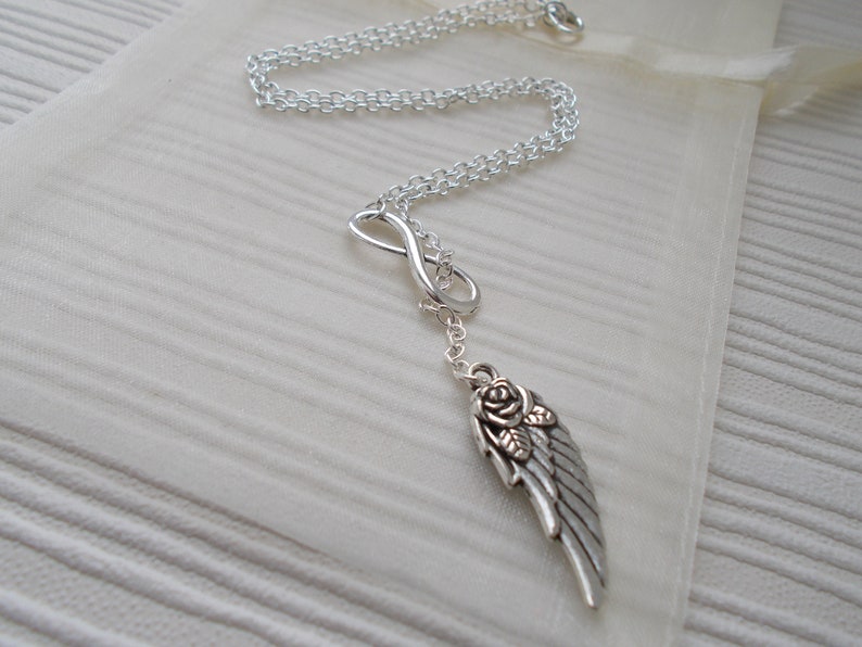 Pu&ograve; includere: Collana color argento con un simbolo dell'infinito e un ciondolo a forma di ala con una rosa. La collana &egrave; esposta su un tessuto color crema, evidenziando il suo design delicato e i dettagli intricati.