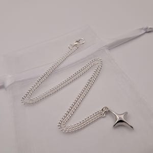 May include: A silver-colored necklace with a four-pointed star pendant. The chain is a curb style, and the pendant has a smooth, polished finish. The necklace is displayed on a white, sheer fabric.