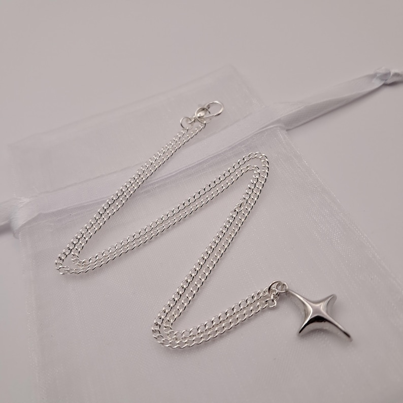 May include: A silver-colored necklace with a four-pointed star pendant. The chain is a curb style, and the pendant has a smooth, polished finish. The necklace is displayed on a white, sheer fabric.