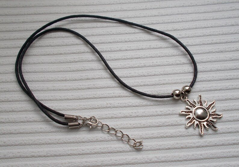 May include: A silver sun pendant necklace with a black cord. The sun pendant has a detailed design with rays extending outwards. The necklace includes a silver clasp and adjustable chain.