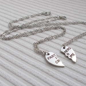 May include: Two silver necklaces with matching heart pendants that say "Partners in Crime" on them. The necklaces are on a white textured surface.