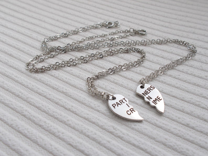 May include: Two silver necklaces with matching heart pendants that say "Partners in Crime" on them. The necklaces are on a white textured surface.