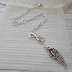 Pu&ograve; includere: Collana color argento con simbolo dell'infinito e ciondolo a forma di ala con dettaglio rosa. La catena &egrave; delicata e la collana &egrave; esposta su un tessuto color crema.