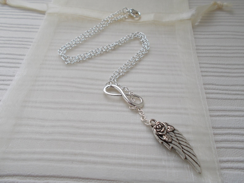 Pu&ograve; includere: Collana color argento con simbolo dell'infinito e ciondolo a forma di ala con dettaglio rosa. La catena &egrave; delicata e la collana &egrave; esposta su un tessuto color crema.