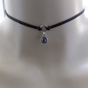 May include: Black choker necklace with a small, round, purple gemstone pendant. The necklace features a thin, black cord and a silver-colored metal ring connecting the pendant. The gemstone is a light purple color.