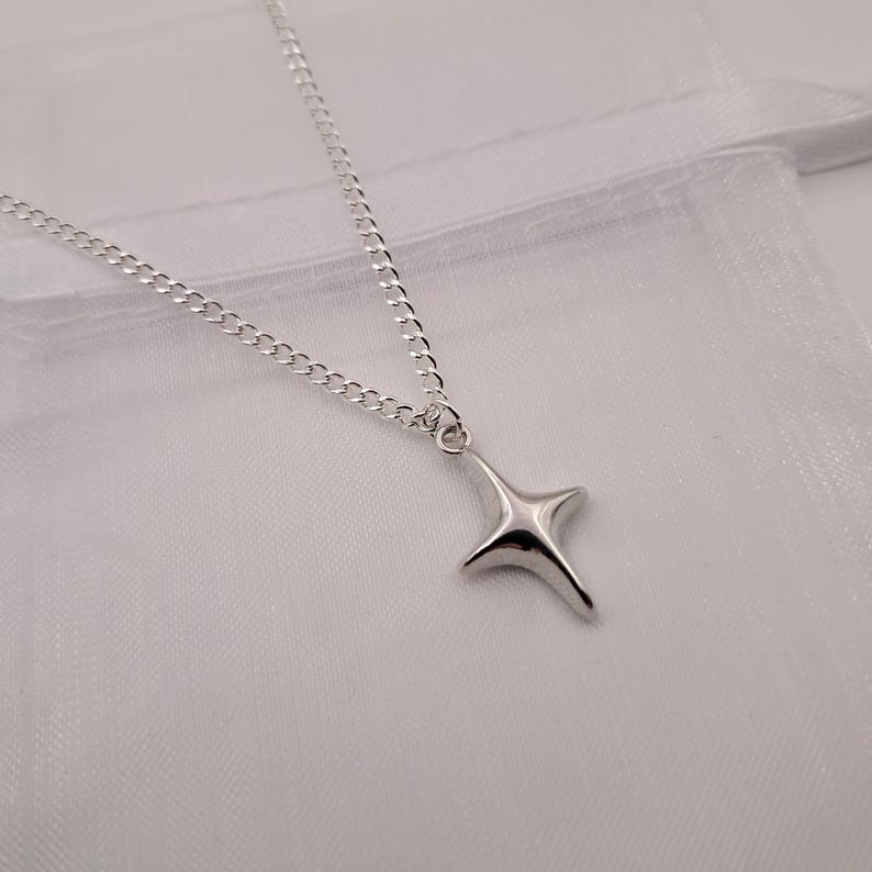 May include: A silver-colored necklace with a four-pointed star pendant. The chain is a delicate curb link style. The pendant has a smooth, reflective surface. The necklace is displayed on a white background.