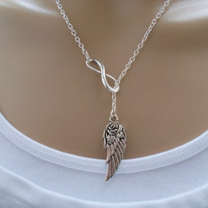 Pu&ograve; includere: Collana color argento con un simbolo dell'infinito e un ciondolo con un design ad ala e rosa. La catena &egrave; delicata e il ciondolo ha un'estetica vintage dettagliata. La collana &egrave; indossata su una maglia bianca.