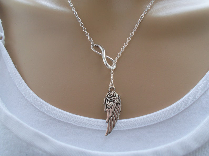 Pu&ograve; includere: Collana color argento con un simbolo dell'infinito e un ciondolo con un design ad ala e rosa. La catena &egrave; delicata e il ciondolo ha un'estetica vintage dettagliata. La collana &egrave; indossata su una maglia bianca.