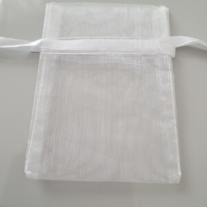 May include: A small, white, sheer organza gift bag with a satin ribbon drawstring closure. The bag is rectangular with a slightly textured appearance. The ribbon is a matching white color.