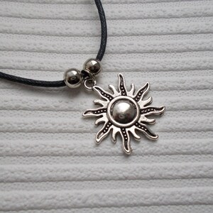 May include: A silver-colored sun pendant necklace with a black cord. The sun pendant has a detailed design with rays and a central circular element. Two silver beads are on the cord near the pendant.