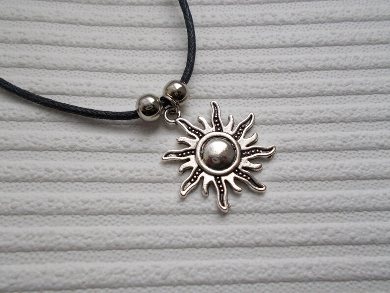 May include: A silver-colored sun pendant necklace with a black cord. The sun pendant has a detailed design with rays and a central circular element. Two silver beads are on the cord near the pendant.