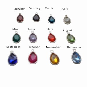May include: A collection of twelve teardrop-shaped pendants, each with a different colored gemstone and a silver-colored loop at the top. The months of the year are printed above each pendant, with colors ranging from red to blue and green.