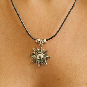 May include: A silver-colored sun pendant necklace with a black cord. The sun pendant has a central sphere surrounded by rays. Two small silver beads are on the cord above the pendant. The necklace is worn around the neck.