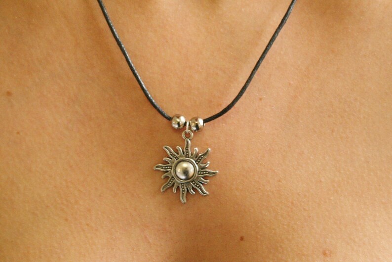 May include: A silver-colored sun pendant necklace with a black cord. The sun pendant has a central sphere surrounded by rays. Two small silver beads are on the cord above the pendant. The necklace is worn around the neck.