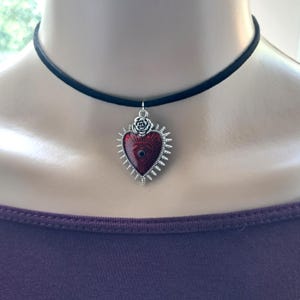 May include: A black choker necklace with a heart-shaped pendant. The pendant is red with a silver-colored eye design and a silver-colored rose at the top. The pendant is surrounded by silver-colored spikes.