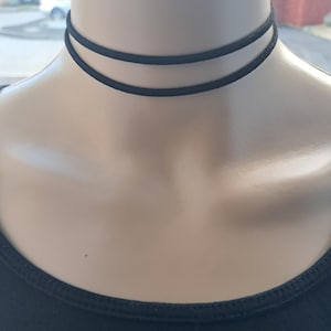 May include: A black double-strand choker necklace featuring two thin, smooth, rounded cords that wrap around the neck. The necklace is worn against a neutral-toned mannequin base, highlighting the simple, minimalist design of the black cord jewelry.