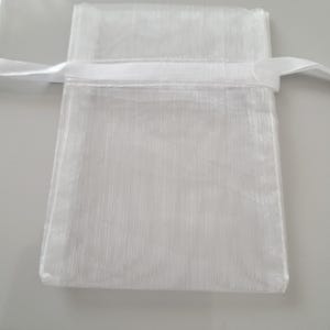 May include: A small, white, sheer fabric gift bag with a satin ribbon drawstring closure. The bag is rectangular with a slightly textured surface. The ribbon is white and tied in a simple knot.