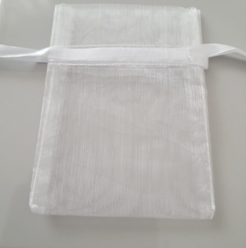 May include: A small, white, sheer fabric gift bag with a satin ribbon drawstring closure. The bag is rectangular with a slightly textured surface. The ribbon is white and tied in a simple knot.