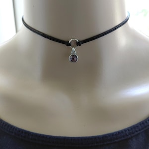 May include: Black choker necklace with a small, round, purple gemstone pendant. The necklace features a thin, black cord and a silver-colored ring connecting the pendant to the cord. The gemstone is a light purple color.