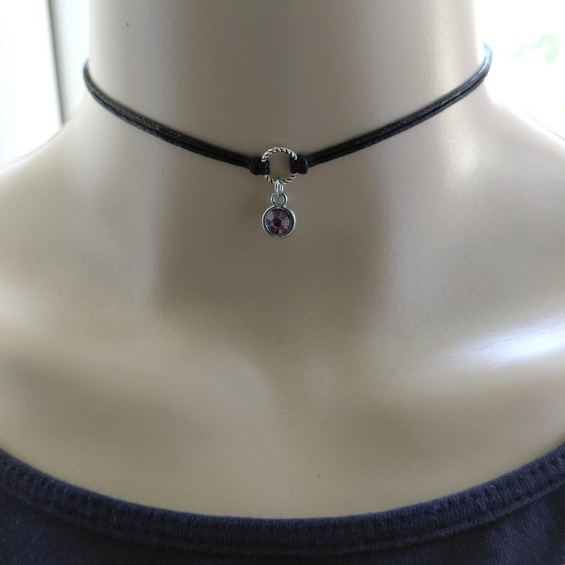 May include: Black choker necklace with a small, round, purple gemstone pendant. The necklace features a thin, black cord and a silver-colored ring connecting the pendant to the cord. The gemstone is a light purple color.