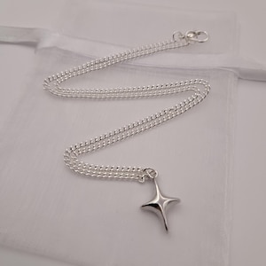 May include: A silver-colored necklace with a four-pointed star pendant. The chain is a delicate curb style, and the pendant is smooth and polished. The necklace is displayed on a white, sheer fabric.