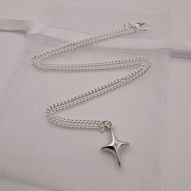 May include: A silver-colored necklace with a four-pointed star pendant. The chain is a delicate curb style, and the pendant is smooth and polished. The necklace is displayed on a white, sheer fabric.