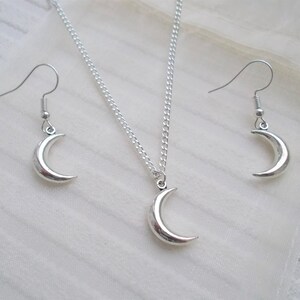 Pu&ograve; includere: Un set di gioielli color argento con una collana con pendente a forma di mezzaluna e orecchini abbinati. La collana ha una catena delicata e gli orecchini hanno chiusure a gancio. Il set &egrave; esposto su una superficie bianca e strutturata.