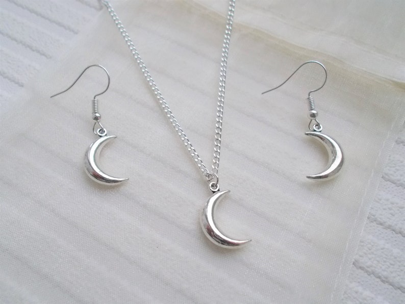 Pu&ograve; includere: Un set di gioielli color argento con una collana con pendente a forma di mezzaluna e orecchini abbinati. La collana ha una catena delicata e gli orecchini hanno chiusure a gancio. Il set &egrave; esposto su una superficie bianca e strutturata.