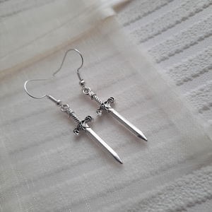May include: A pair of silver sword earrings with a detailed design. The earrings are hanging from silver hooks.