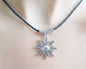 May include: A silver sun pendant necklace with a black leather cord. The pendant has a detailed sun design with a round center.