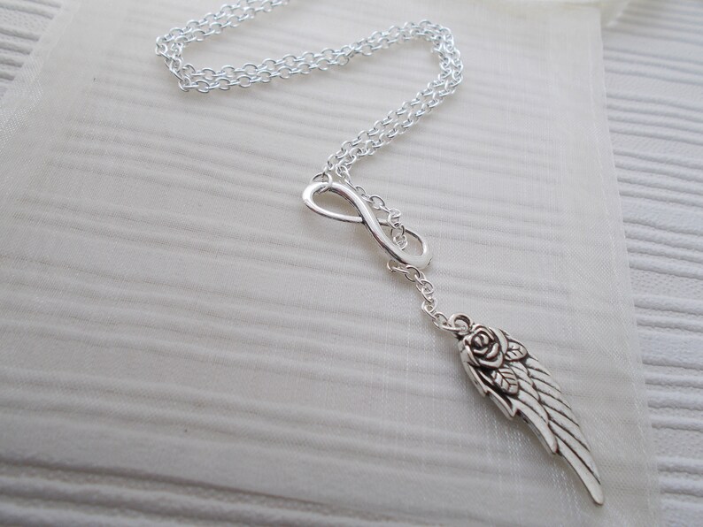 Pu&ograve; includere: Collana color argento con un simbolo dell'infinito e un ciondolo a forma di ala con un dettaglio a rosa. La catena &egrave; delicata e il ciondolo &egrave; finemente lavorato. La collana &egrave; esposta su una superficie bianca e strutturata.