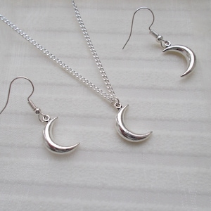 Pu&ograve; includere: Un set di gioielli color argento con una collana con pendente a mezzaluna e orecchini abbinati. La collana ha una catena delicata e gli orecchini hanno chiusure a gancio. Il set &egrave; esposto su una superficie bianca e strutturata.