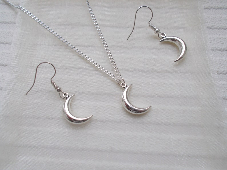 Pu&ograve; includere: Un set di gioielli color argento con una collana con pendente a mezzaluna e orecchini abbinati. La collana ha una catena delicata e gli orecchini hanno chiusure a gancio. Il set &egrave; esposto su una superficie bianca e strutturata.