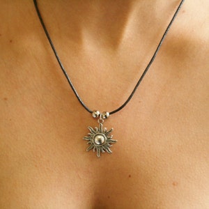 May include: A silver sun pendant necklace with a black cord. The sun pendant has a detailed design with a central sphere and radiating rays. The necklace is worn around the neck.
