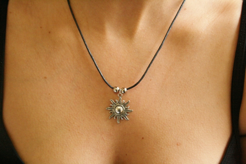 May include: A silver sun pendant necklace with a black cord. The sun pendant has a detailed design with a central sphere and radiating rays. The necklace is worn around the neck.