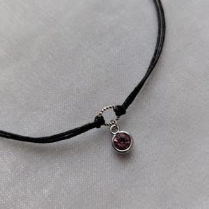 May include: A delicate black cord necklace featuring a small, round, pink gemstone pendant. The pendant is set in a silver-colored metal. The necklace is displayed on a white, textured surface.