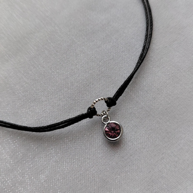May include: A delicate black cord necklace featuring a small, round, pink gemstone pendant. The pendant is set in a silver-colored metal. The necklace is displayed on a white, textured surface.
