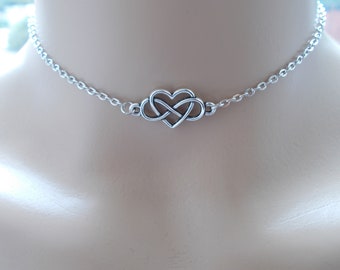 May include: A silver-toned metal choker necklace featuring a central charm that combines a heart shape with an infinity symbol. The charm is connected to a delicate, thin link chain that rests around the neck, showcasing a polished metallic finish.