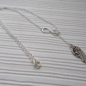 Pu&ograve; includere: Una collana color argento con un simbolo dell'infinito e un ciondolo a forma di ala con un dettaglio a rosa. La collana ha una catena delicata e una chiusura a moschettone. Il gioiello &egrave; esposto su una superficie bianca.