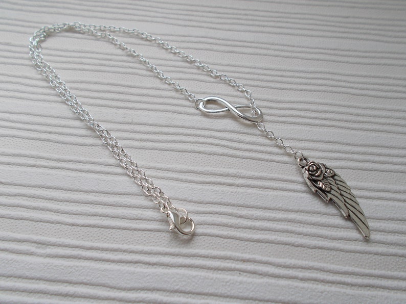 Pu&ograve; includere: Una collana color argento con un simbolo dell'infinito e un ciondolo a forma di ala con un dettaglio a rosa. La collana ha una catena delicata e una chiusura a moschettone. Il gioiello &egrave; esposto su una superficie bianca.
