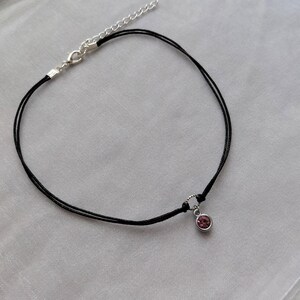 May include: A delicate black choker necklace with a small, round, pink gemstone pendant. The necklace features a silver-toned clasp and adjustable chain. The pendant is set in a silver-toned bezel.