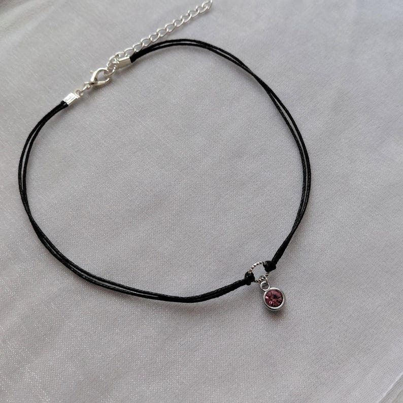 May include: A delicate black choker necklace with a small, round, pink gemstone pendant. The necklace features a silver-toned clasp and adjustable chain. The pendant is set in a silver-toned bezel.