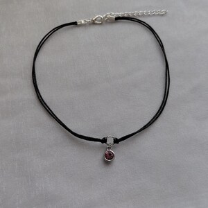 May include: A black choker necklace with a small, round, pink gemstone pendant. The necklace has a silver-colored clasp and an adjustable chain. The choker is made of two thin black cords.