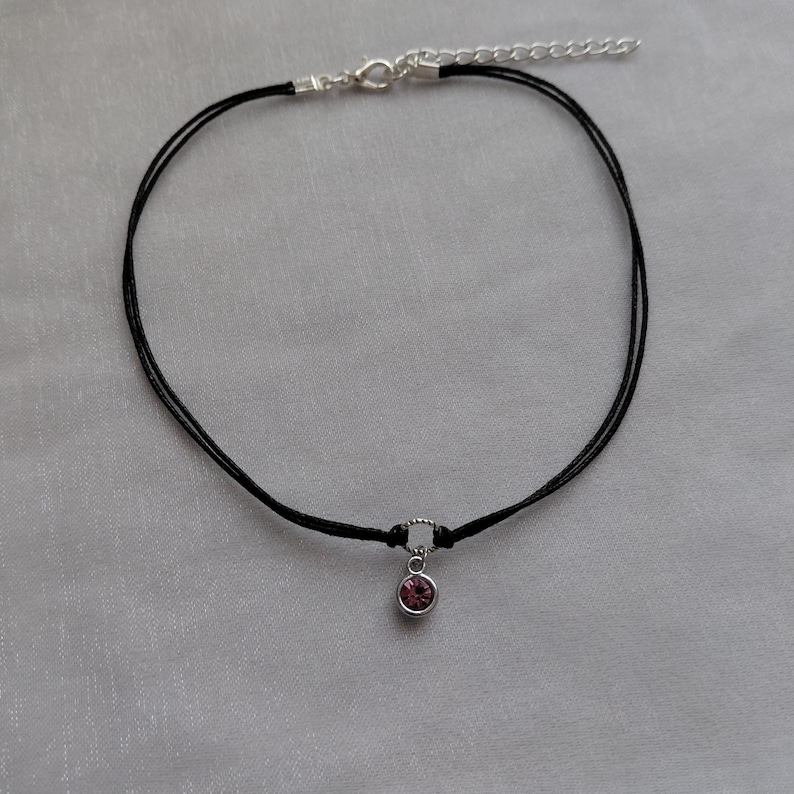May include: A black choker necklace with a small, round, pink gemstone pendant. The necklace has a silver-colored clasp and an adjustable chain. The choker is made of two thin black cords.