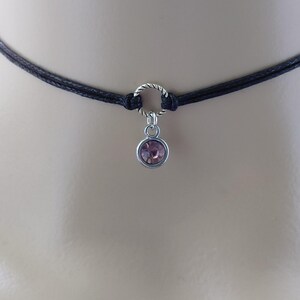 May include: A black choker necklace with a silver-colored ring and a small, round, pink gemstone pendant. The necklace is made of two strands of black cord. The pendant is a light pink color and is set in a silver-colored metal.