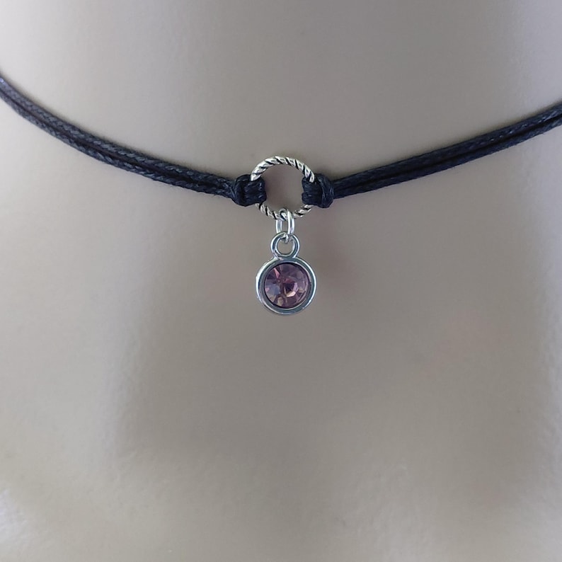 May include: A black choker necklace with a silver-colored ring and a small, round, pink gemstone pendant. The necklace is made of two strands of black cord. The pendant is a light pink color and is set in a silver-colored metal.