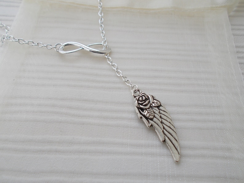Pu&ograve; includere: Collana color argento con un simbolo dell'infinito e un ciondolo con un'ala e un design a rosa. La collana ha una catena delicata ed &egrave; esposta su uno sfondo morbido e neutro.