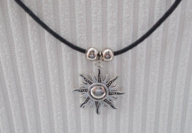 May include: A silver-colored sun pendant necklace with a black cord. The sun pendant has a detailed design with rays and a central circular element. Two silver beads are on the cord above the pendant.