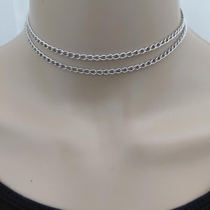 May include: A double-layered silver-toned chain choker necklace featuring two delicate, interconnected curb chains that rest closely around the neck. The metallic links have a polished finish, creating a simple, layered appearance that sits flush against the skin.