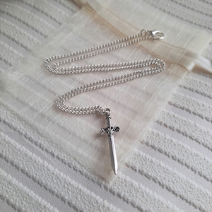 May include: A silver chain necklace with a silver sword pendant. The sword has a detailed design and a pointed tip.